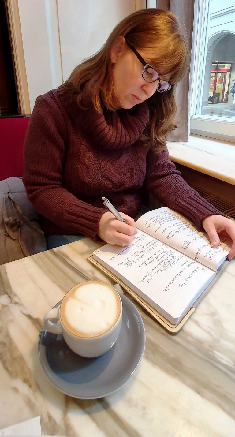 Constanze Scheib in einem Kaffeehaus, schreibt in ein Notizbuch, vor ihr eine Tasse Kaffee