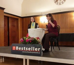 Buchhändlerin Julia Jessenitschnig und Autorin Constanze Scheib an einem Tisch. Constanze Scheib mit einem Mikrofon in der einen Hand, mit der anderen gestikulierend