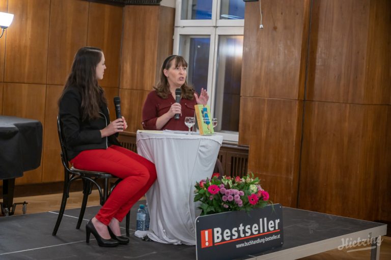 Buchhändlerin Julia Jessenitschnig und Autorin Constanze Scheib mit Mikrofonen in der Hand auf einer Bühne an einem Tisch sitzend. Auf dem Tisch ein Exemplar von "Der Würger von Hietzing" davor ein Aufsteller: "Buchhandlung Bestseller"